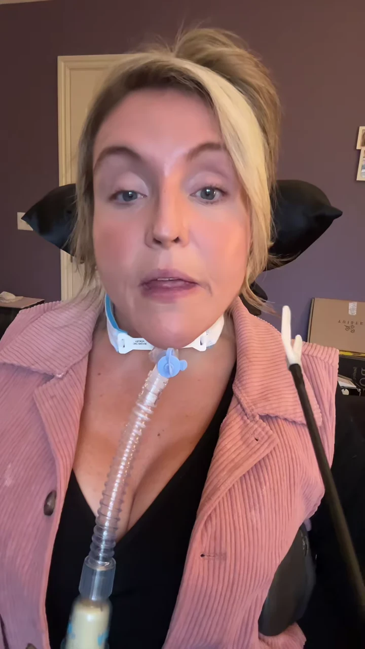 Laura Michelle wearing a pink jacket and looking into the camera with her tracheostomy tubing visible.